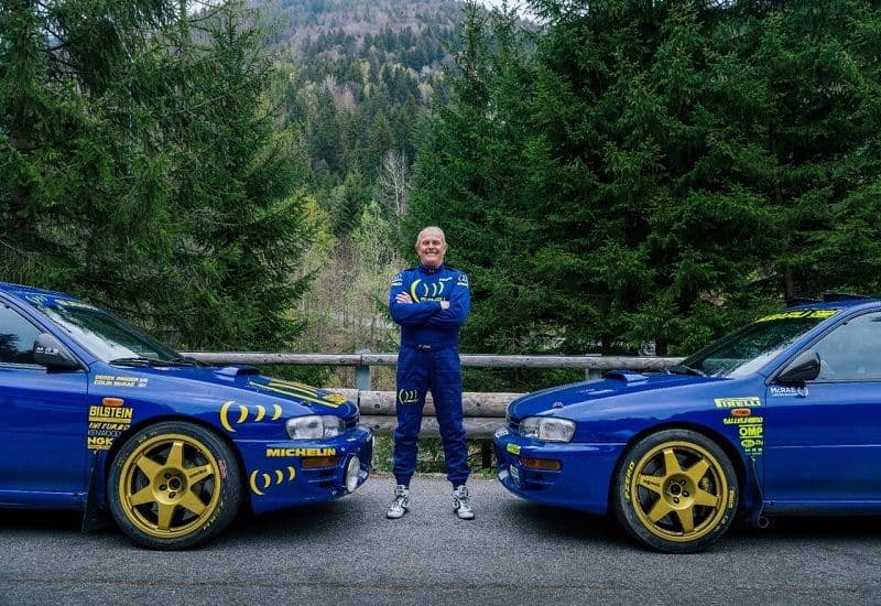 Pilota in tuta blu sorridente tra due Subaru Impreza da rally blu con cerchi dorati.