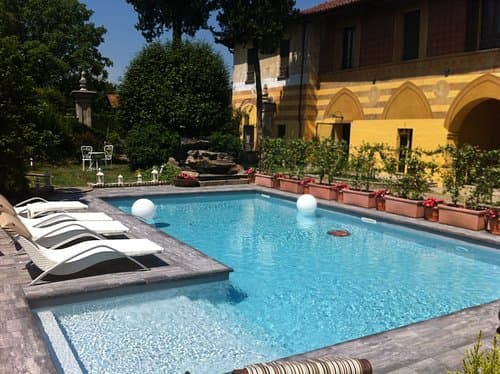 Piscina azzurra in un giardino con lettini bianchi e una villa gialla sullo sfondo.